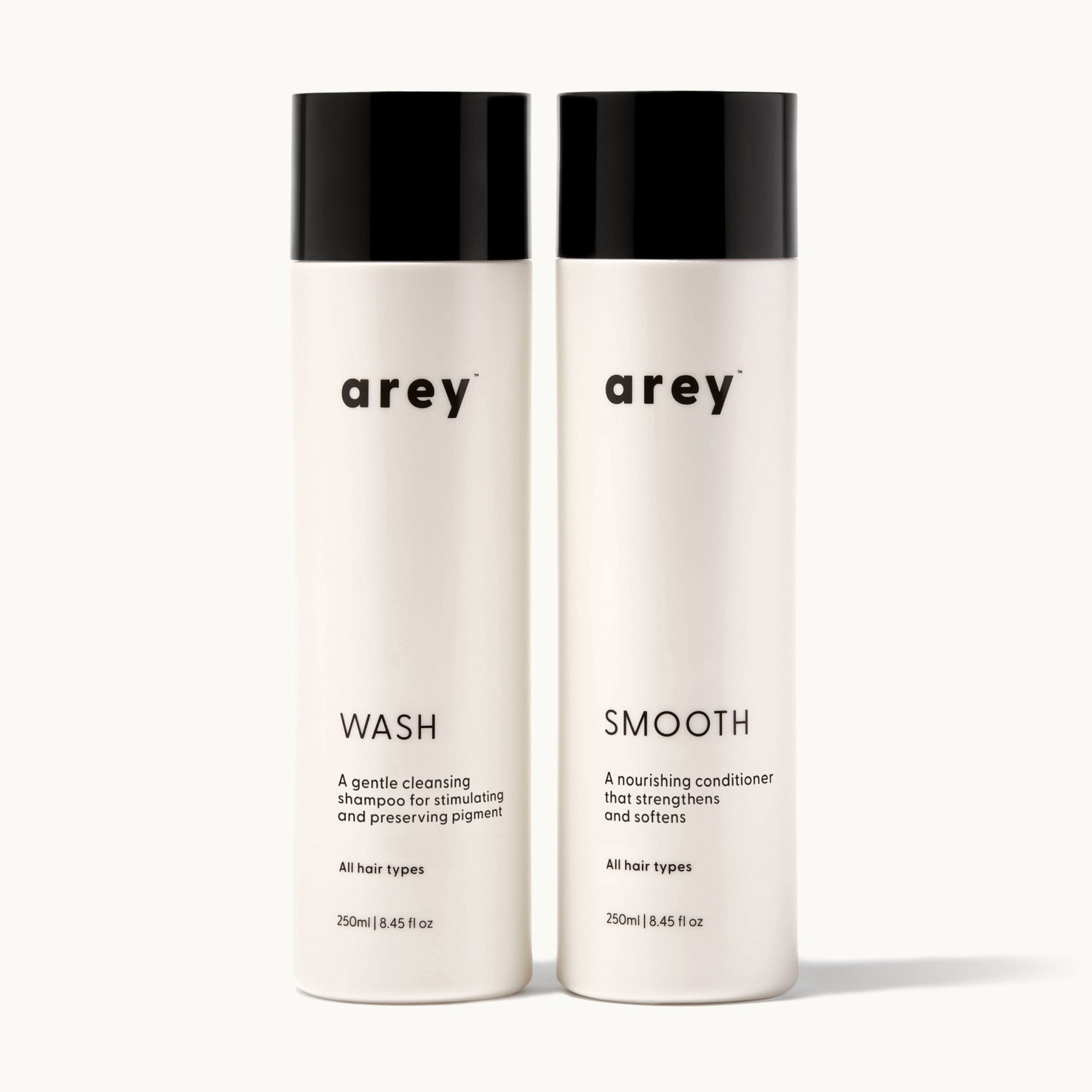 Arey Shower Duo on cream background. Two bottles side by side: Wash (A gentle cleansing shampoo for stimulating and preserving pigment. All hair types. 250ml / 8.45 fl oz) and Smooth (A nourishing conditioner that strengthens and softens. All hair types. 250ml / 8.45 fl oz).