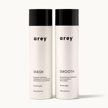 Arey Shower Duo on cream background. Two bottles side by side: Wash (A gentle cleansing shampoo for stimulating and preserving pigment. All hair types. 250ml / 8.45 fl oz) and Smooth (A nourishing conditioner that strengthens and softens. All hair types. 250ml / 8.45 fl oz).