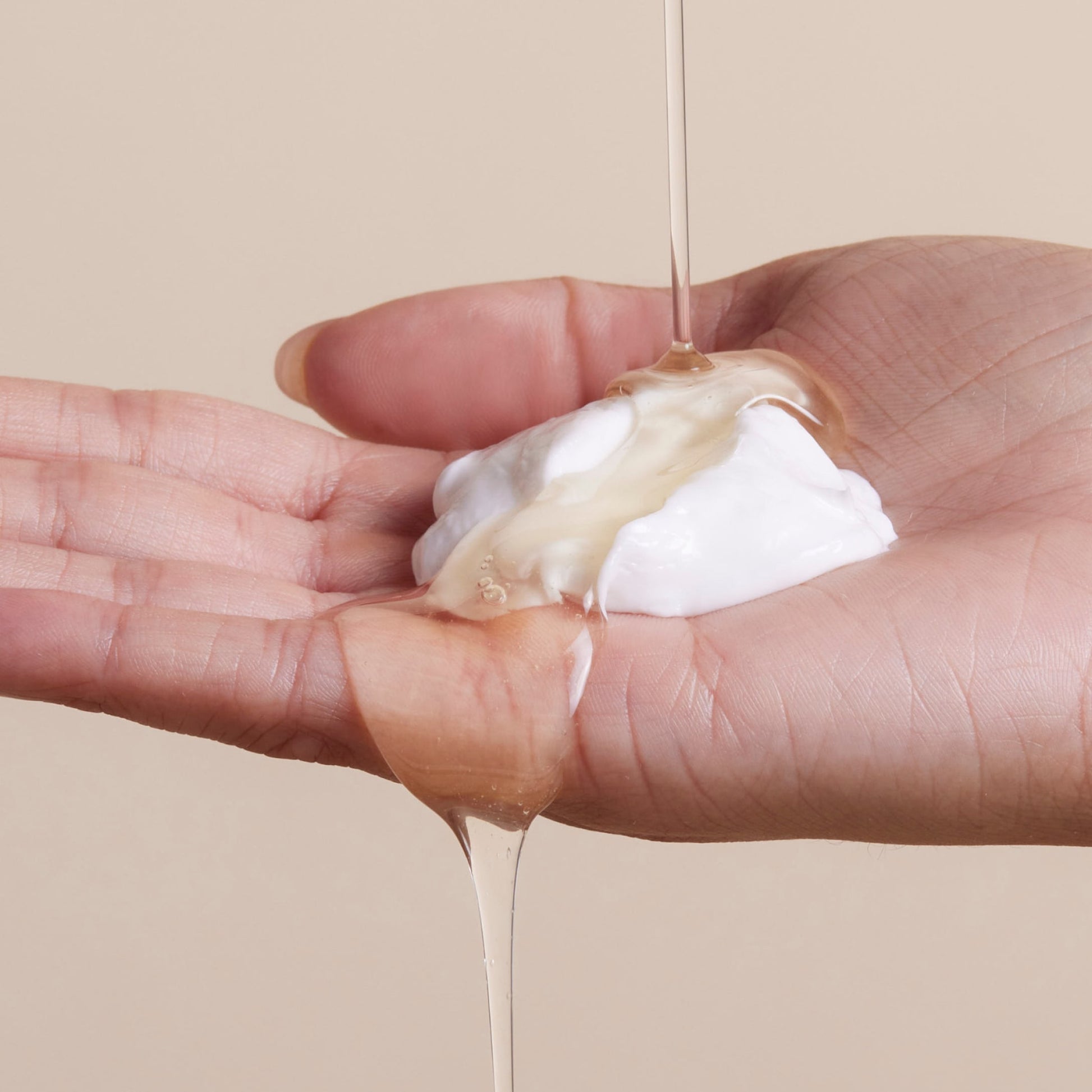 A hand holds a dollop of white cream as a clear, viscous liquid from Arey’s The Shower Duo is poured over it against a neutral beige background—evoking the rejuvenating in-shower hair care ritual that cleanses and restores for healthier hair.