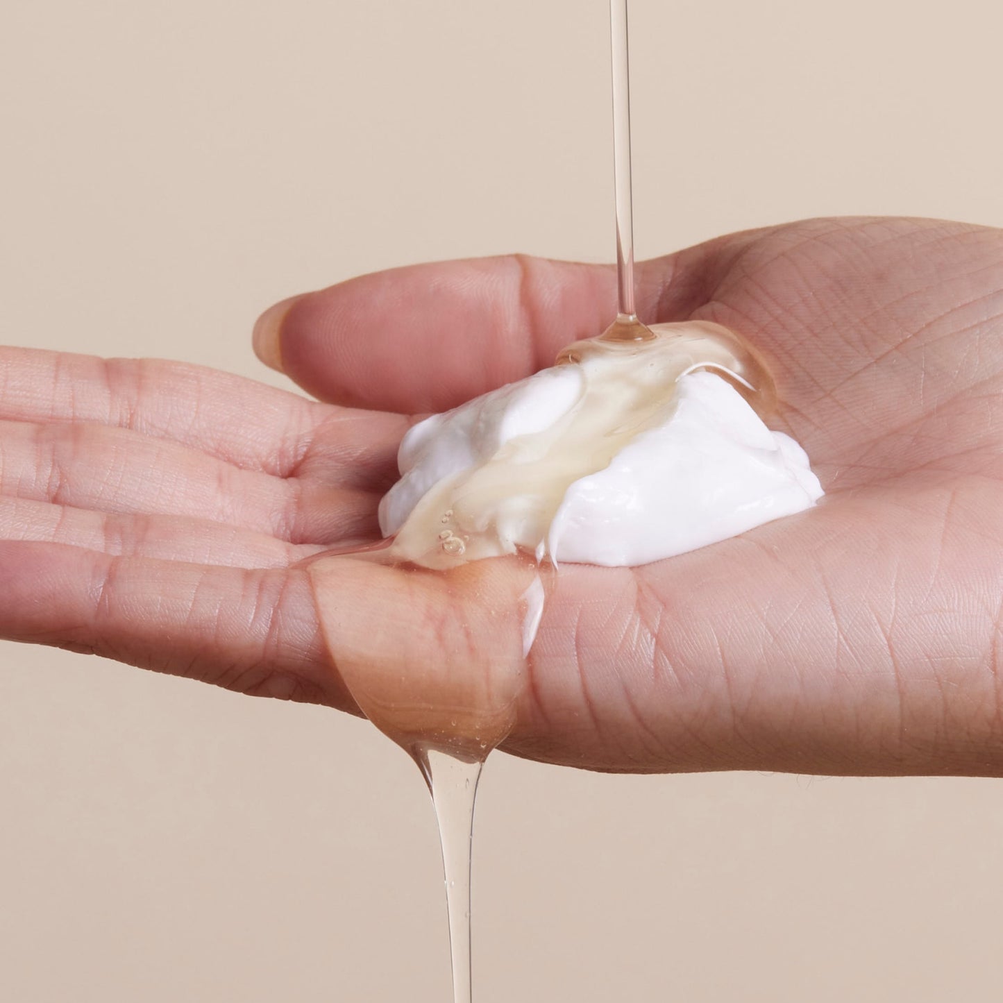 A hand holds a dollop of white cream as a clear, viscous liquid from Arey’s The Shower Duo is poured over it against a neutral beige background—evoking the rejuvenating in-shower hair care ritual that cleanses and restores for healthier hair.