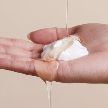 A hand holds a dollop of white cream as a clear, viscous liquid from Arey’s The Shower Duo is poured over it against a neutral beige background—evoking the rejuvenating in-shower hair care ritual that cleanses and restores for healthier hair.