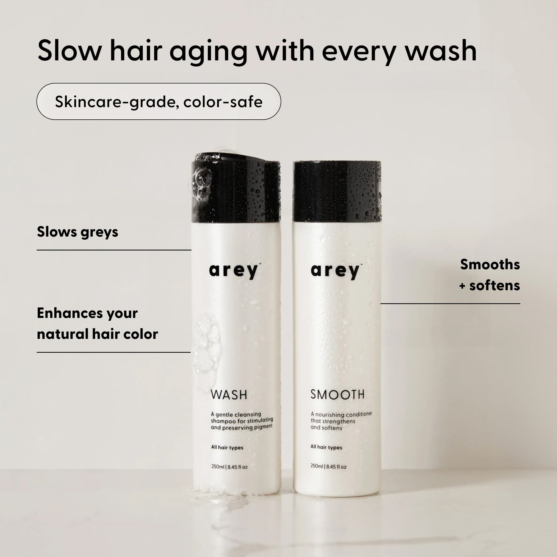 Two Arey The Shower Duo bottles labeled Wash and Smooth stand on a white surface. Text highlights the benefits: slows greys, enhances your natural hair color, and smooths + softens for healthier, more vibrant hair. Experience in-shower care from Arey that cleanses and restores your hair’s natural radiance.