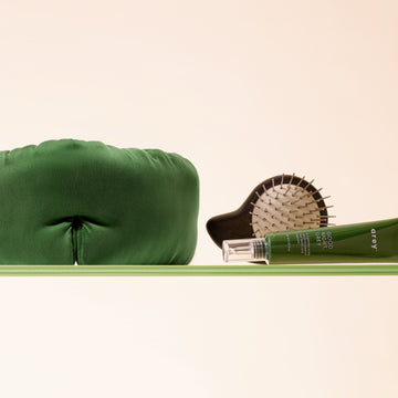 The Sleep Ritual Set by Arey, featuring a green sleep mask, a scalp brush with white bristles, and a green tube of overnight scalp serum, is arranged neatly on a light green surface against a beige background, creating an inviting setup for hair wellness.