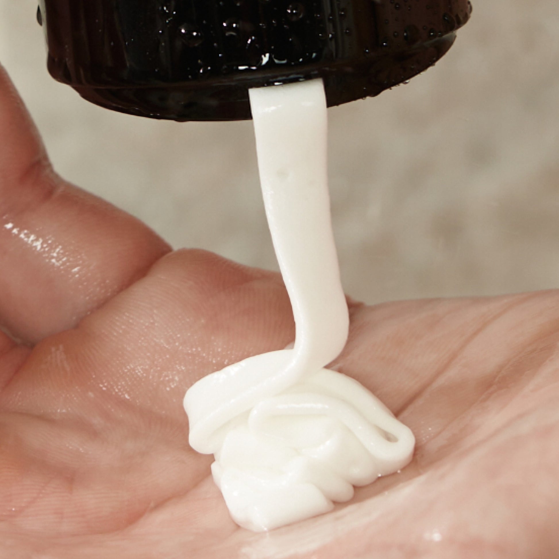 A close-up of Arey’s Smooth Conditioner, a white nourishing cream, being squeezed from a black bottle onto the palm of a person’s hand.