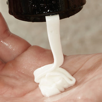 A close-up of Arey’s Smooth Conditioner, a white nourishing cream, being squeezed from a black bottle onto the palm of a person’s hand.