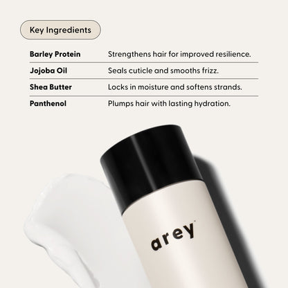 A bottle labeled Arey Smooth Conditioner lies next to a smear of cream. Text lists key ingredients—barley protein, jojoba oil, shea butter, and panthenol—and highlights their benefits for hair health and hydration. This nourishing conditioner from Arey uses a clean formula to leave your hair feeling smoother, healthier, and deeply hydrated.