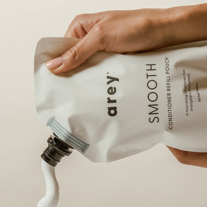 A hand squeezes Arey Smooth Conditioner Refill from a white pouch, dispensing white cream into an unseen container—a nourishing conditioner designed to repair and care for hair.
