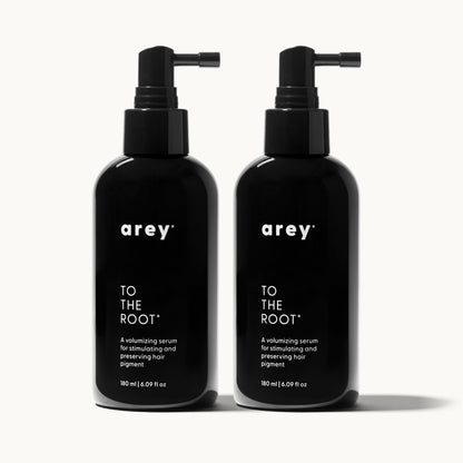 Two Arey To The Root serum bottles on a cream background, representing a 6-month supply. Both bottles labeled: arey. TO THE ROOT. A volumizing serum for stimulating and preserving hair pigment. 180 ml / 6.09 fl oz.