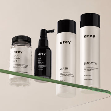 Four Arey hair care products displayed on a glass shelf against a neutral background. Left to right: Not Today, Grey dietary supplement (clear jar), To The Root scalp serum (black spray bottle, 66ml), Wash shampoo (white bottle, 250ml), and Smooth conditioner (white bottle, 250ml).