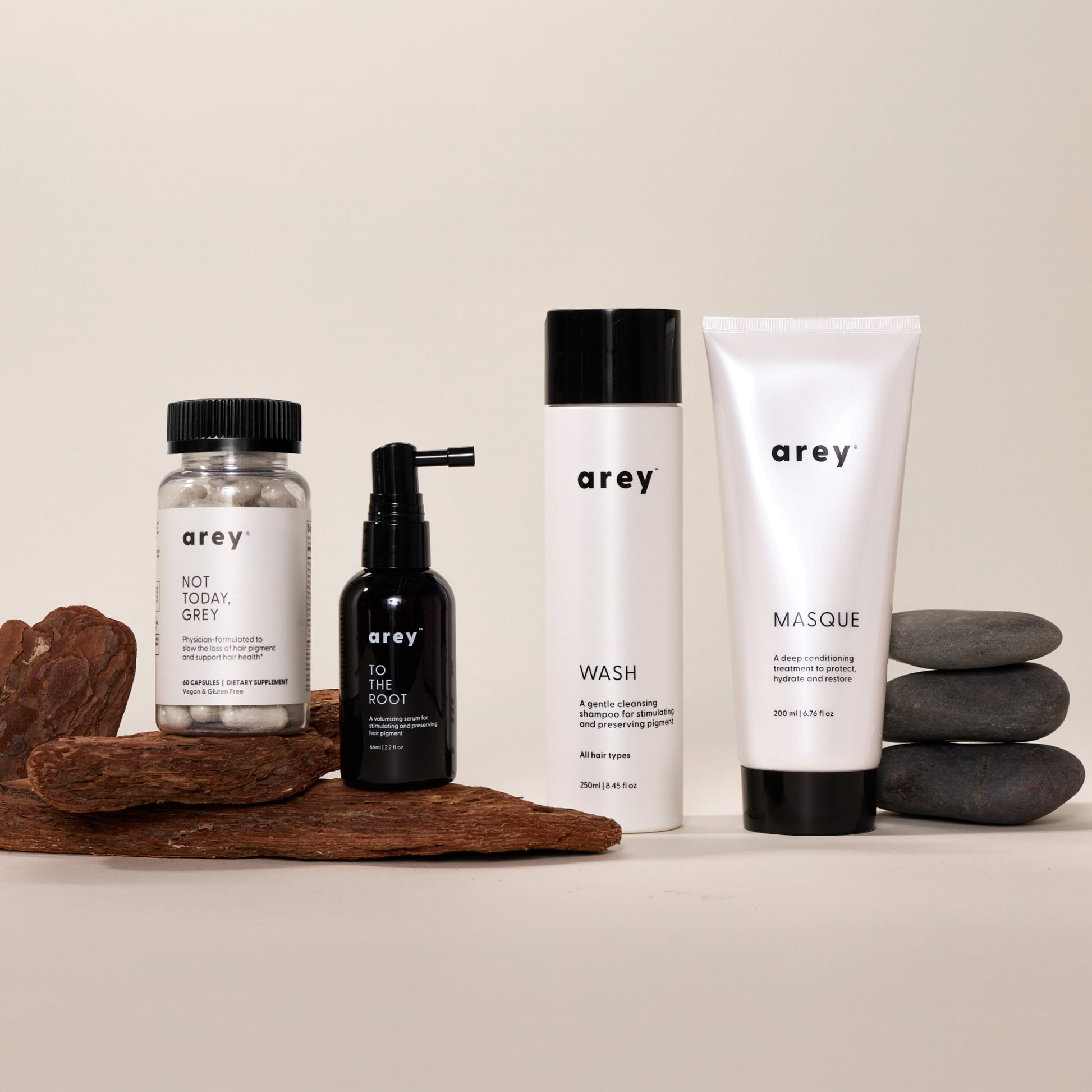 Four Arey products from The Restorative Kit—Not Today, Grey pills, To The Root serum, Wash shampoo, and Masque—are displayed on a surface with wood pieces and stacked smooth stones against a neutral background. Developed by Arey to support repigmentation and slow grey hair growth, The Restorative Kit combines advanced science with natural elements for healthier-looking hair.