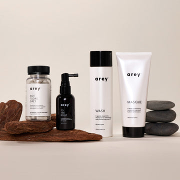 Four Arey products from The Restorative Kit—Not Today, Grey pills, To The Root serum, Wash shampoo, and Masque—are displayed on a surface with wood pieces and stacked smooth stones against a neutral background. Developed by Arey to support repigmentation and slow grey hair growth, The Restorative Kit combines advanced science with natural elements for healthier-looking hair.