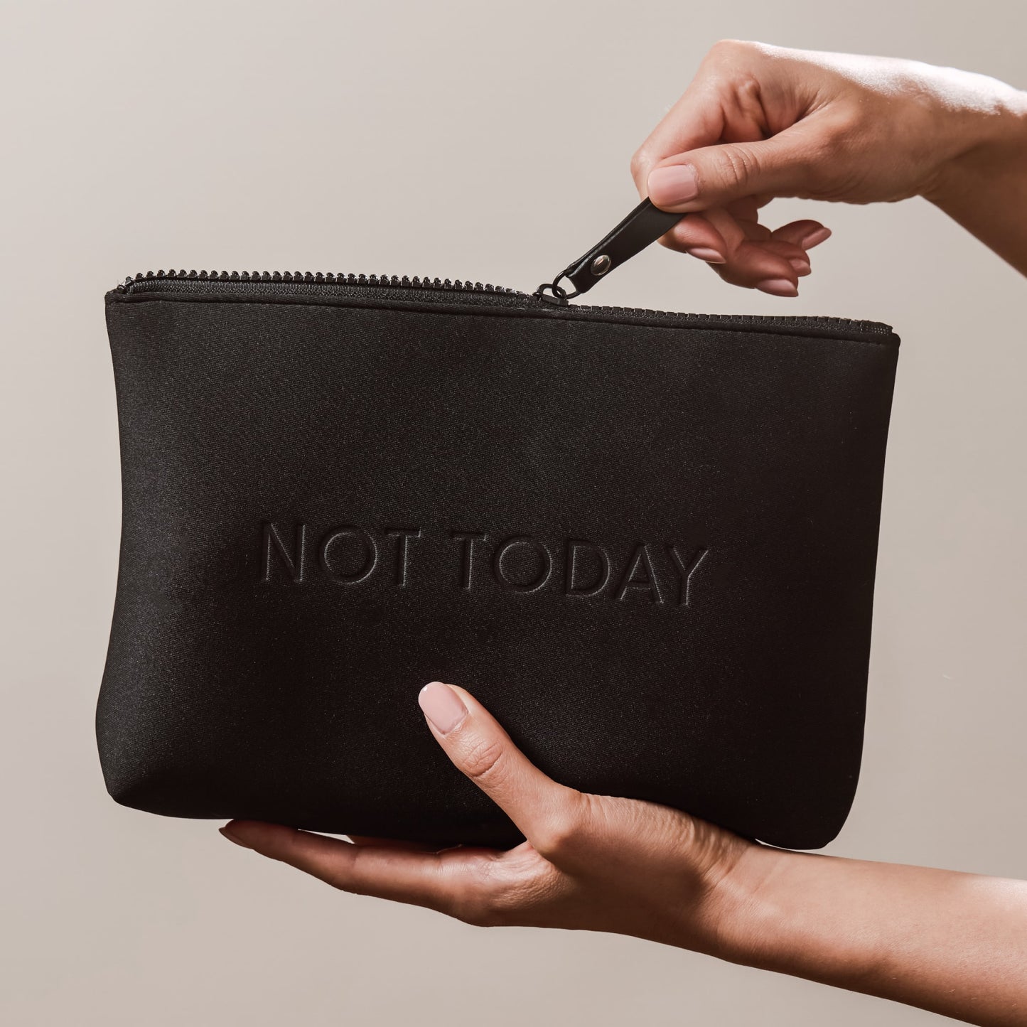 Hands holding the Arey "Not Today" black travel bag. One hand unzips the top while the other supports the bottom. The bag has "NOT TODAY" embossed on the front.