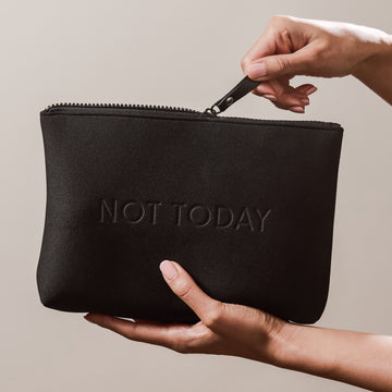 Hands holding the Arey "Not Today" black travel bag. One hand unzips the top while the other supports the bottom. The bag has "NOT TODAY" embossed on the front.