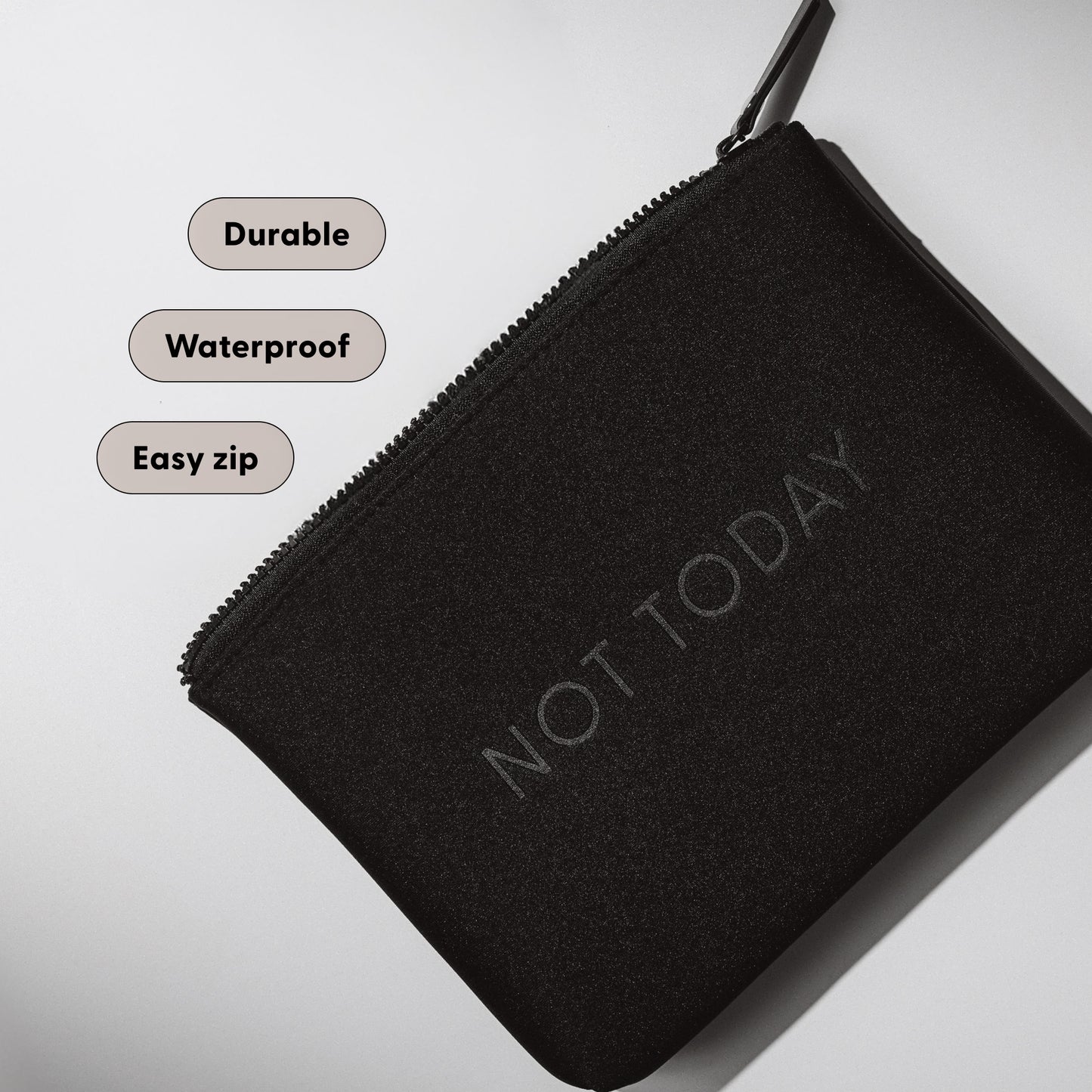 Close-up of the Arey "Not Today" black travel bag with feature labels. The zippered pouch has "NOT TODAY" embossed on it. Labels read: Durable. Waterproof. Easy zip.