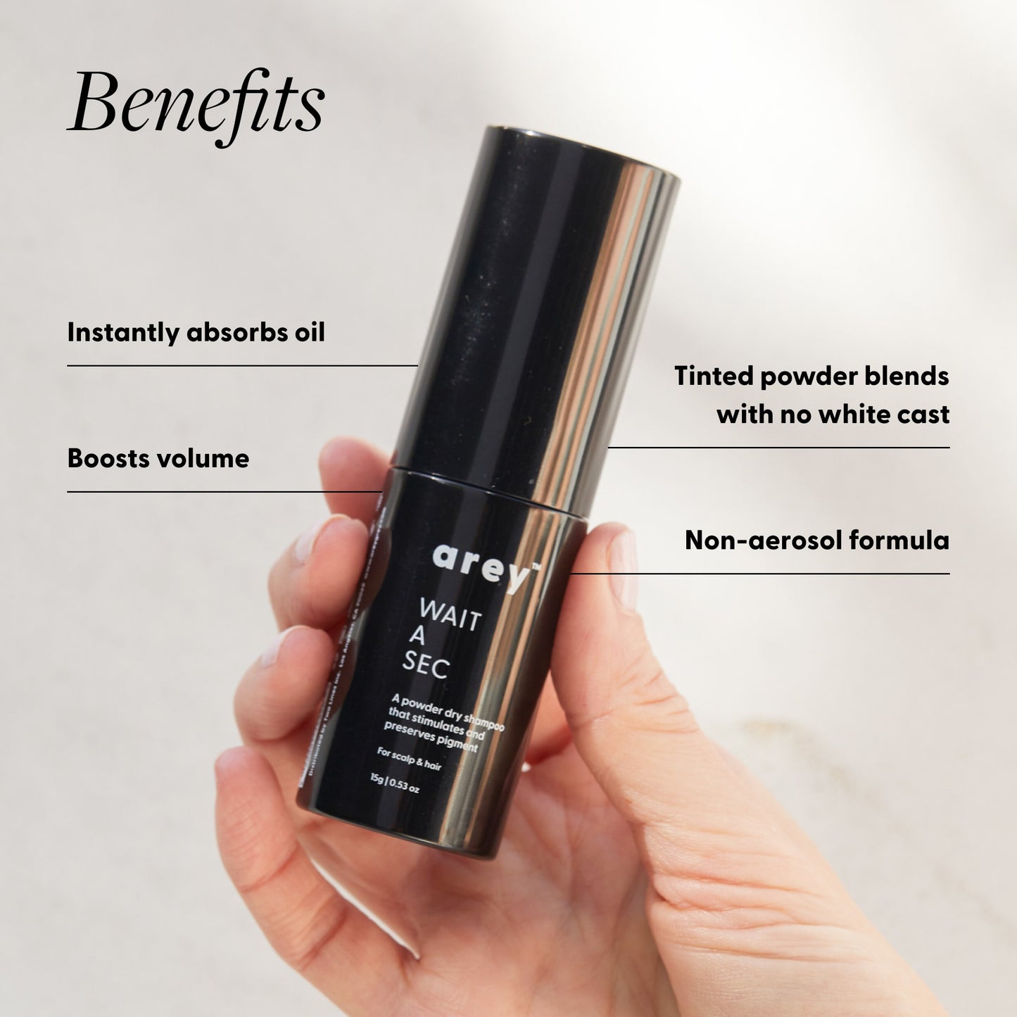 Hand holding Arey Wait A Sec bottle with benefits labeled. Text reads: Benefits - Instantly absorbs oil. Tinted powder blends with no white cast. Boosts volume. Non-aerosol formula. Bottle label reads: arey. WAIT A SEC. A powder dry shampoo that stimulates and preserves pigment. For scalp & hair. 15g / 0.53 oz.