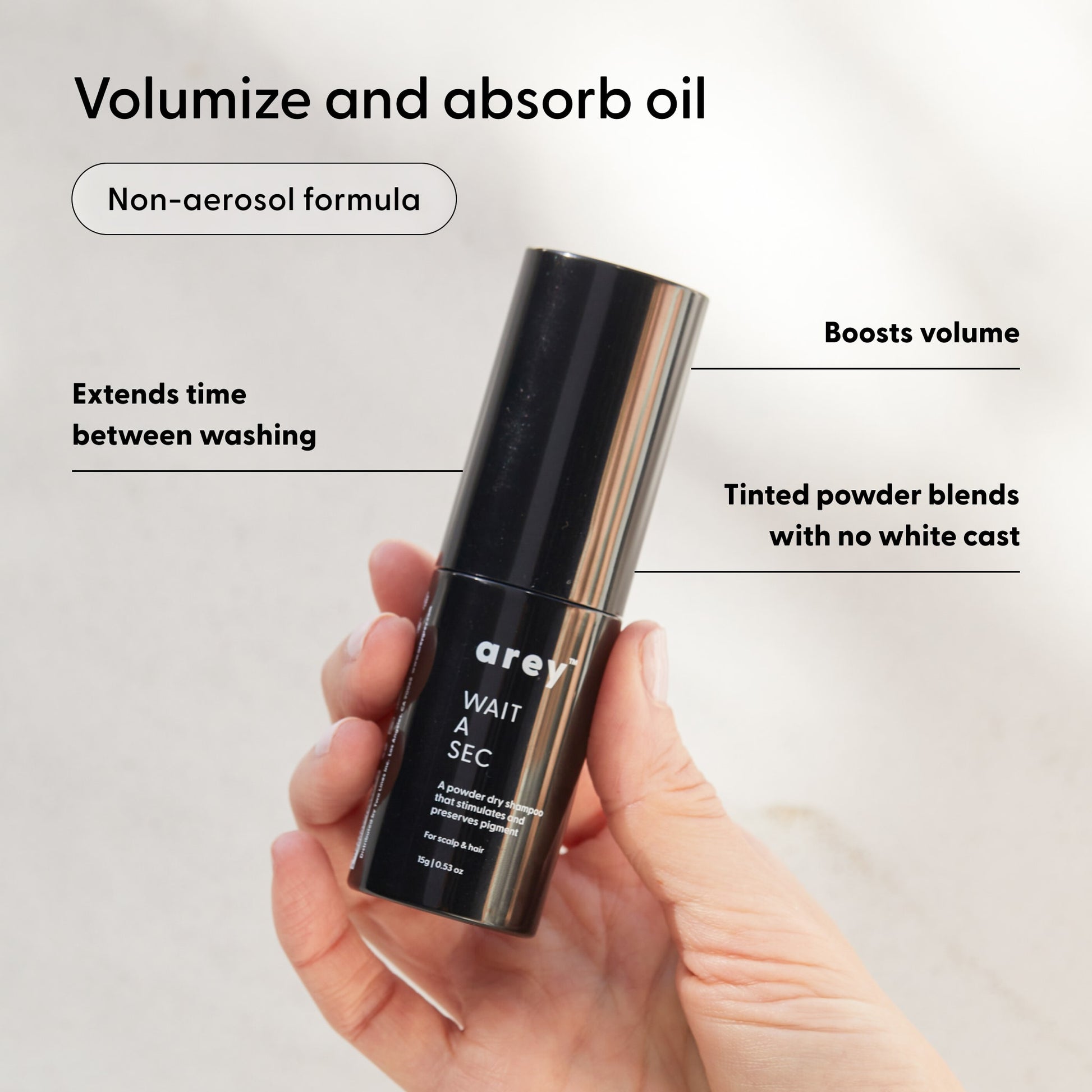 A hand holds a black bottle labeled “Arey Wait A Sec® Dry Shampoo.” Text around the image highlights its features: a non-aerosol formula that absorbs oil, boosts volume, extends time between washes, and uses tinted powder that blends seamlessly with no white cast.