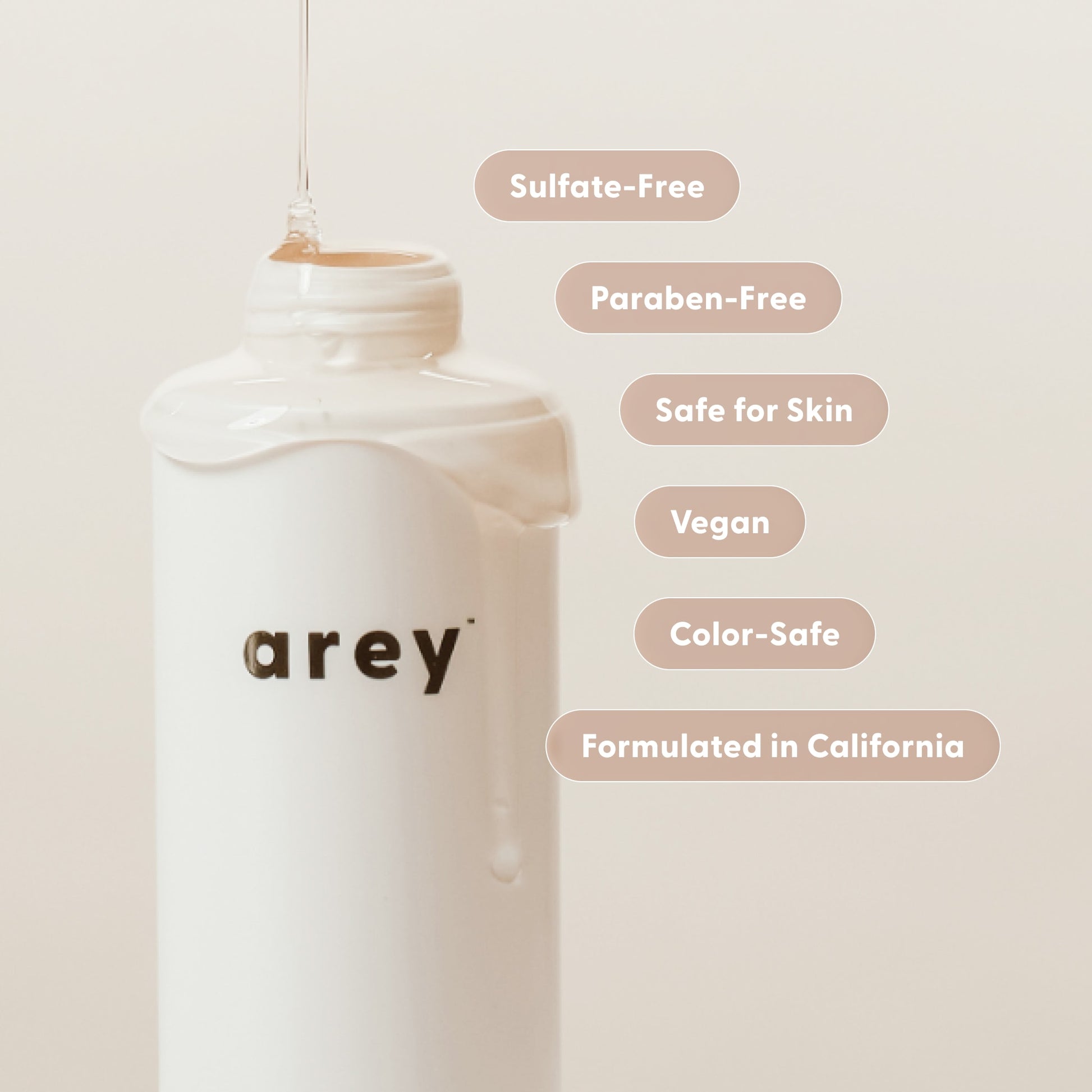 A white bottle labeled Arey Wash Shampoo is featured against a beige background, with liquid pouring onto it. Text bubbles showcase benefits such as Sulfate-Free, Paraben-Free, Safe for Skin, Vegan, Color-Safe, and Formulated in California. This peptide-based shampoo by Arey is expertly formulated to promote hair health and gently care for gray strands while providing anti-aging benefits.