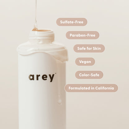 A white bottle labeled Arey Wash Shampoo is featured against a beige background, with liquid pouring onto it. Text bubbles showcase benefits such as Sulfate-Free, Paraben-Free, Safe for Skin, Vegan, Color-Safe, and Formulated in California. This peptide-based shampoo by Arey is expertly formulated to promote hair health and gently care for gray strands while providing anti-aging benefits.