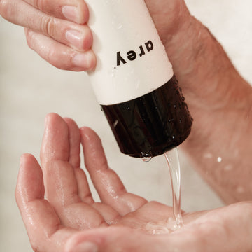 A person dispenses clear liquid from a white bottle labeled Arey Wash Shampoo into their wet palm, with water droplets visible on the bottle and hands, highlighting the refreshing effects of this peptide shampoo.