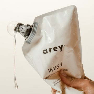 A hand holds a large Arey Wash Shampoo Refill pouch, with shampoo flowing from the nozzle and forming bubbles—an ideal option for those looking for a convenient and hair-healthy way to refill their shampoo.