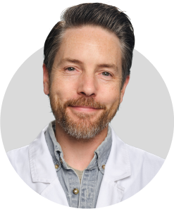 A man with short dark hair, a beard, and mustache smiles at the camera. He is wearing a light denim shirt and a white lab coat, with a plain light gray background.