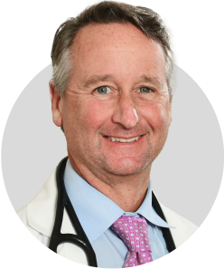 A middle-aged man wearing a white coat, blue shirt, and pink tie smiles at the camera. A stethoscope is draped around his neck. The background is light gray.