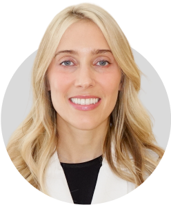Smiling woman with long blonde hair wearing a white coat and black top, pictured against a light gray background.